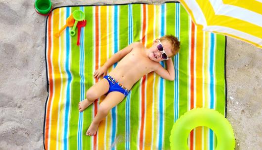 Bambino in spiaggia su asciugamano colorato con occhiali da sole.