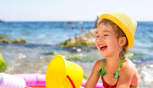 Bambina sorridente con cappello giallo gioca in spiaggia.